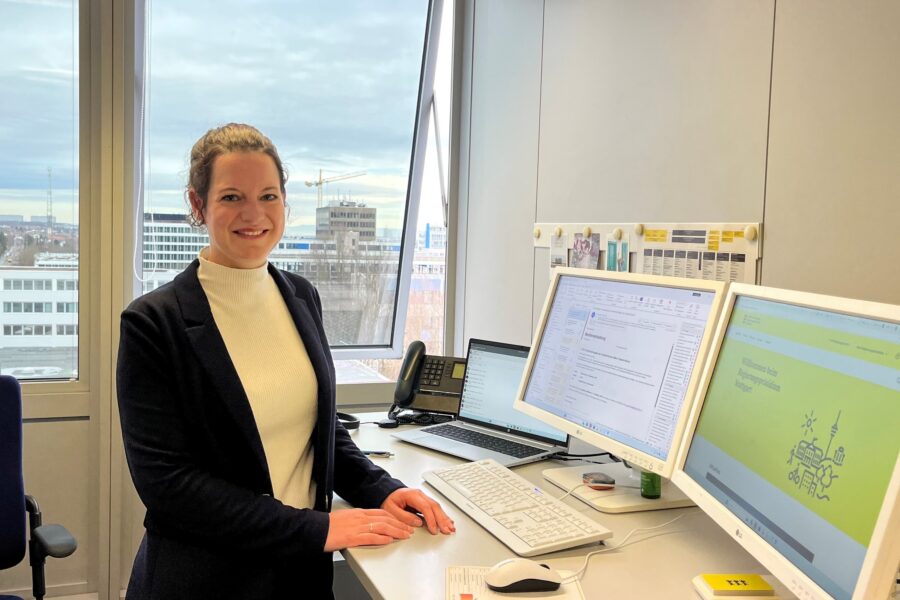 Frau steht lächelnd an einem Schreibtisch mit zwei Monitoren und einem Fenster im Hintergrund.