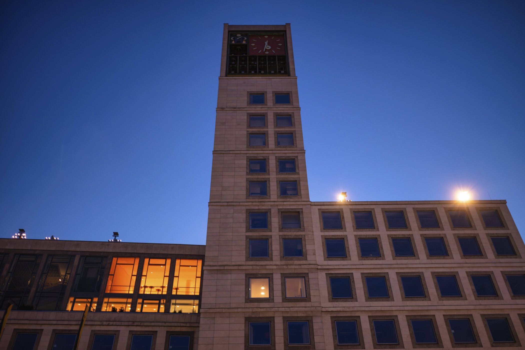 Hohes Gebäude mit Uhrturm, beleuchtete Fenster, blauer Himmel im Hintergrund.