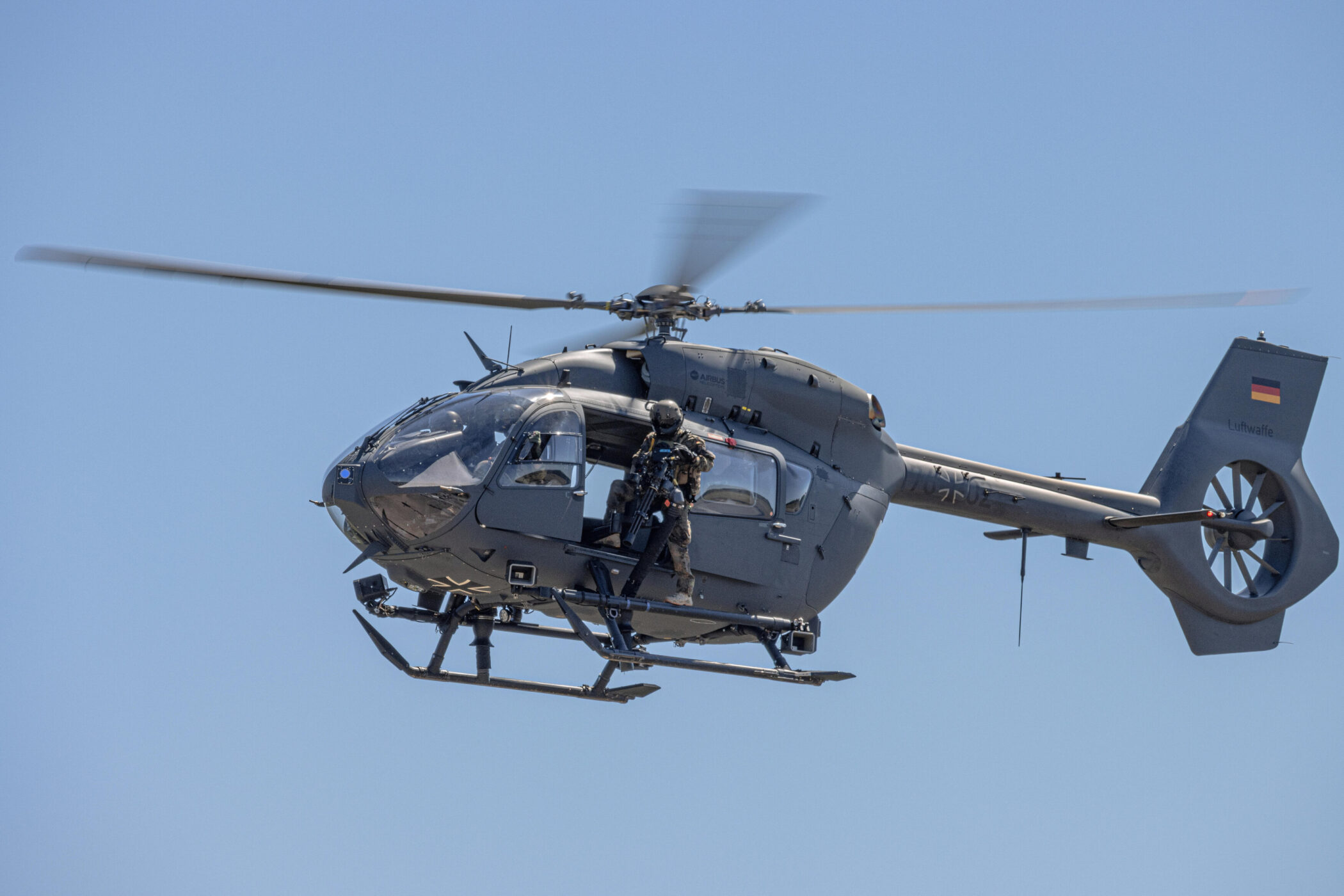 Grauer Hubschrauber mit Soldat in der Tür, blauer Himmel im Hintergrund.