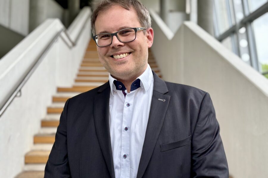 Mann mit Brille und Anzug lächelt vor einer Treppe in einem modernen Gebäude.