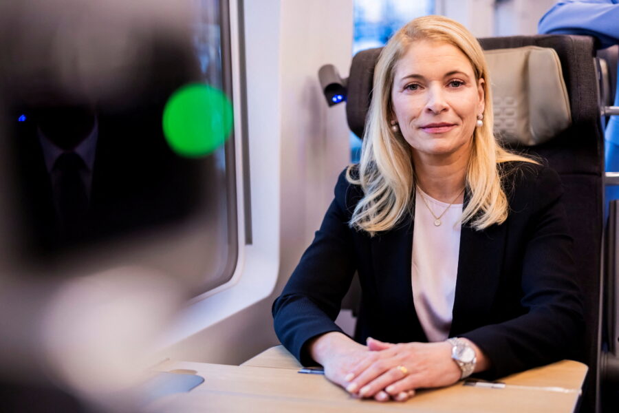 Eine Frau sitzt in einem Zugabteil an einem Tisch und schaut in die Kamera.