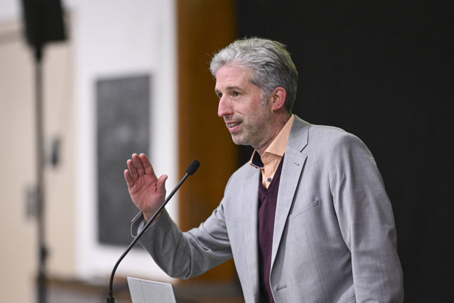 Mann in grauem Anzug spricht in ein Mikrofon, hebt die Hand.