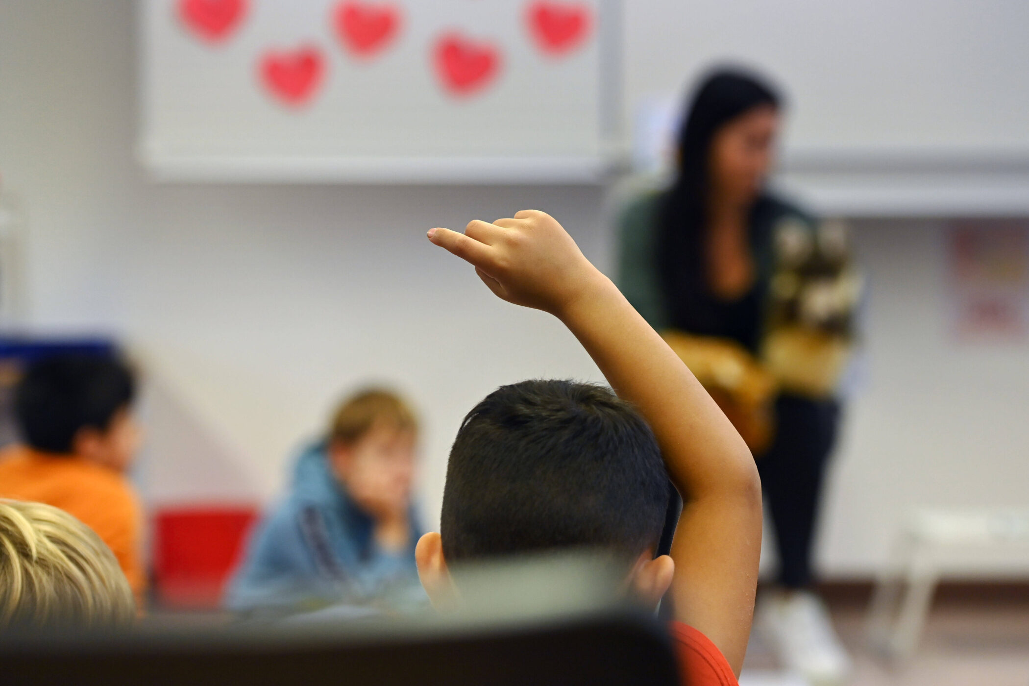 Kind hebt Hand in Klassenzimmer, Lehrerin im Hintergrund.