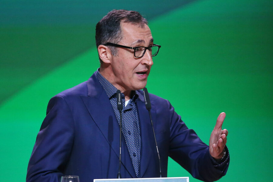 Mann mit Brille spricht gestikulierend vor grünem Hintergrund.