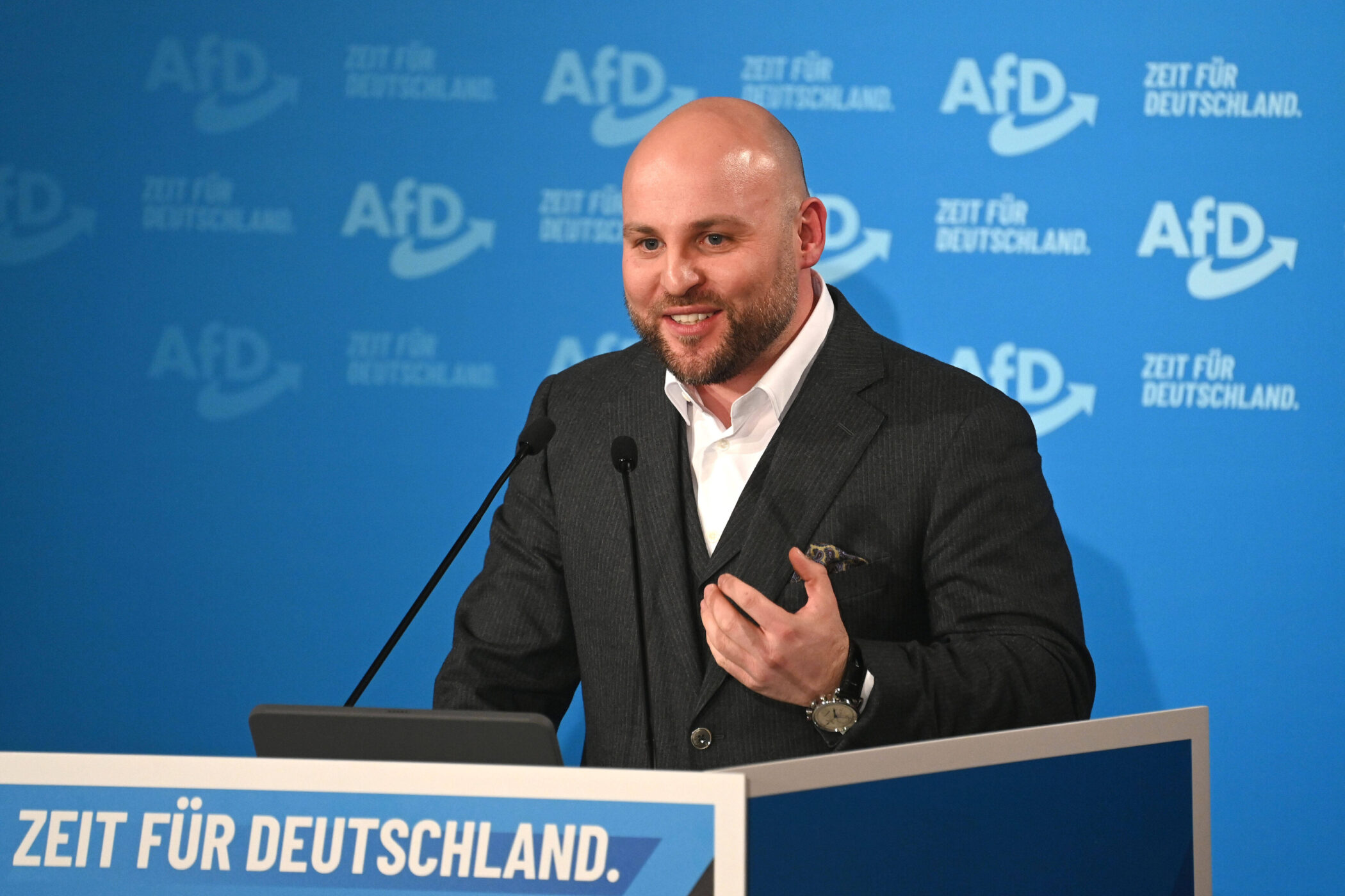 Mann am Rednerpult, Hintergrund mit AfD-Logos und Text "Zeit für Deutschland".