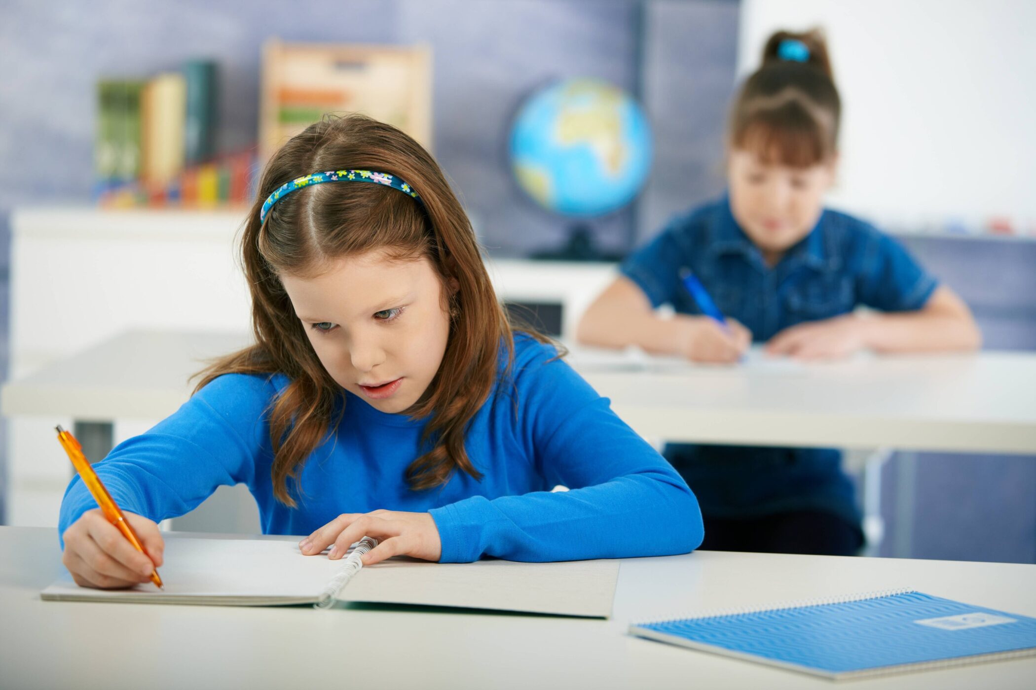 Zwei Kinder sitzen an Tischen und schreiben in Hefte. Im Hintergrund ein Globus.