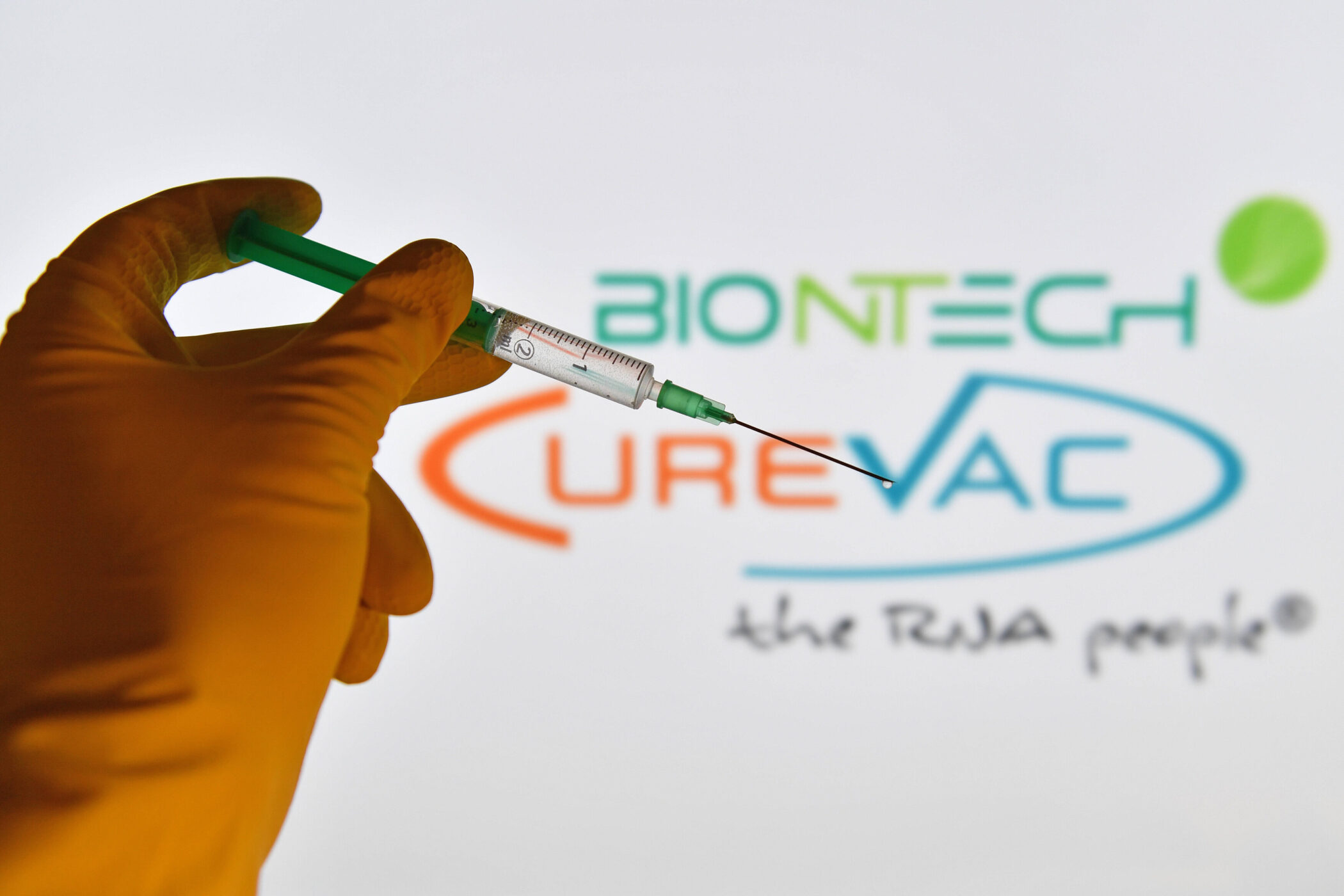 Hand mit Spritze vor unscharfem BioNTech- und CureVac-Logo.