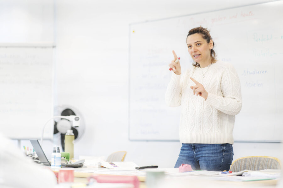 Frau gestikuliert vor einer Tafel in einem Klassenzimmer.