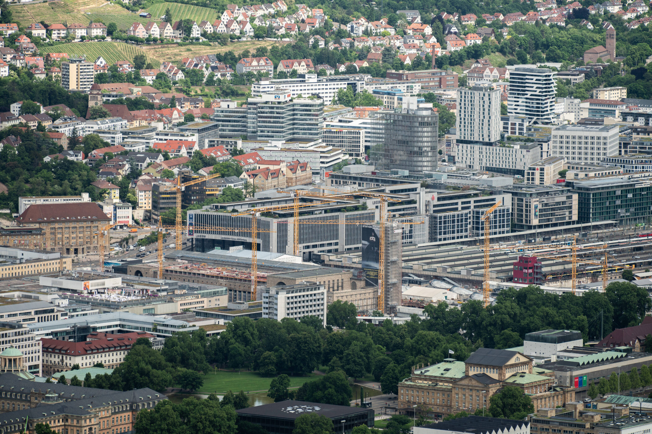 Stadtansicht mit modernen Gebäuden, Baukränen und grüner Umgebung im Hintergrund.