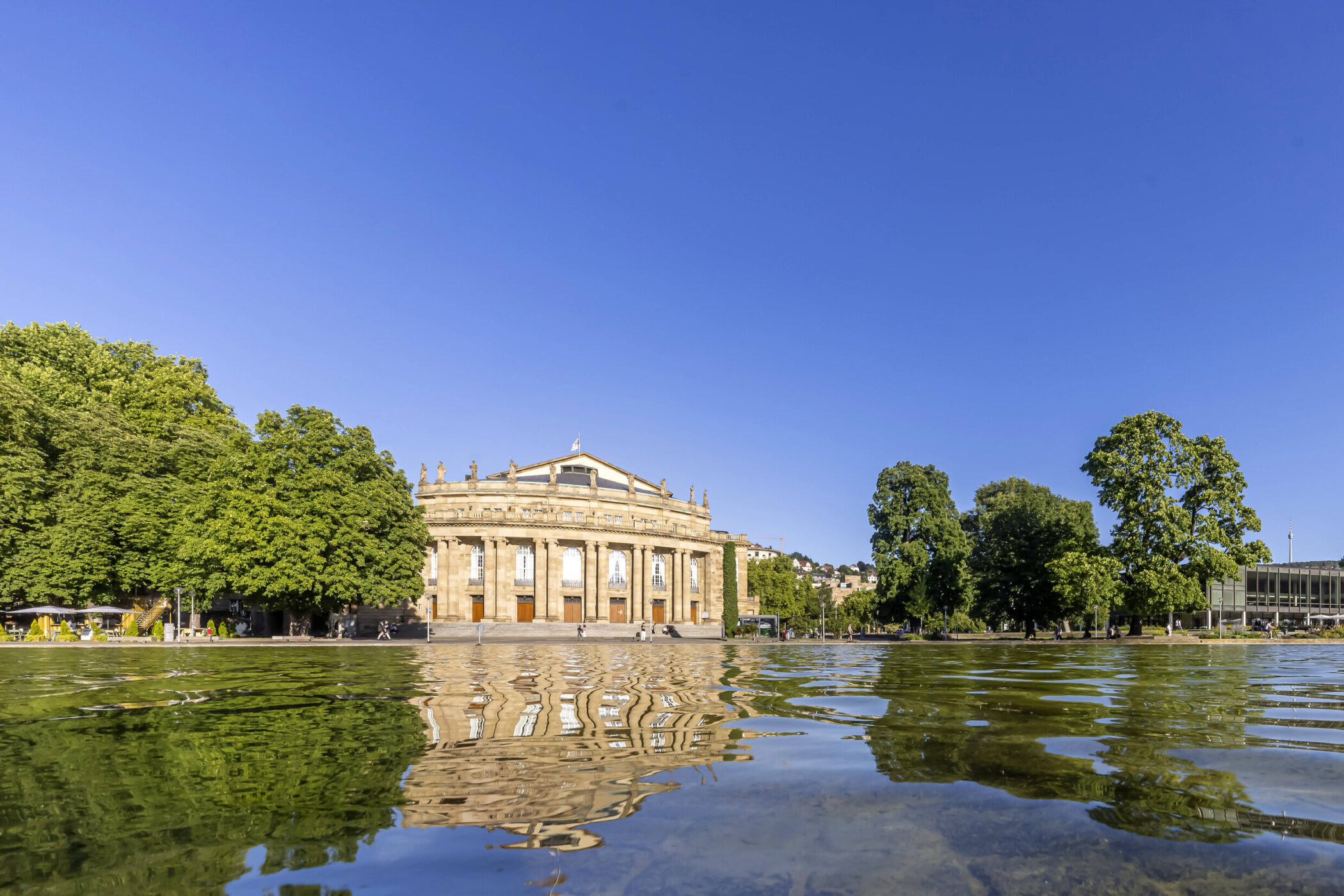 Gebäude mit Säulen hinter einem Teich, umgeben von Bäumen, blauer Himmel.