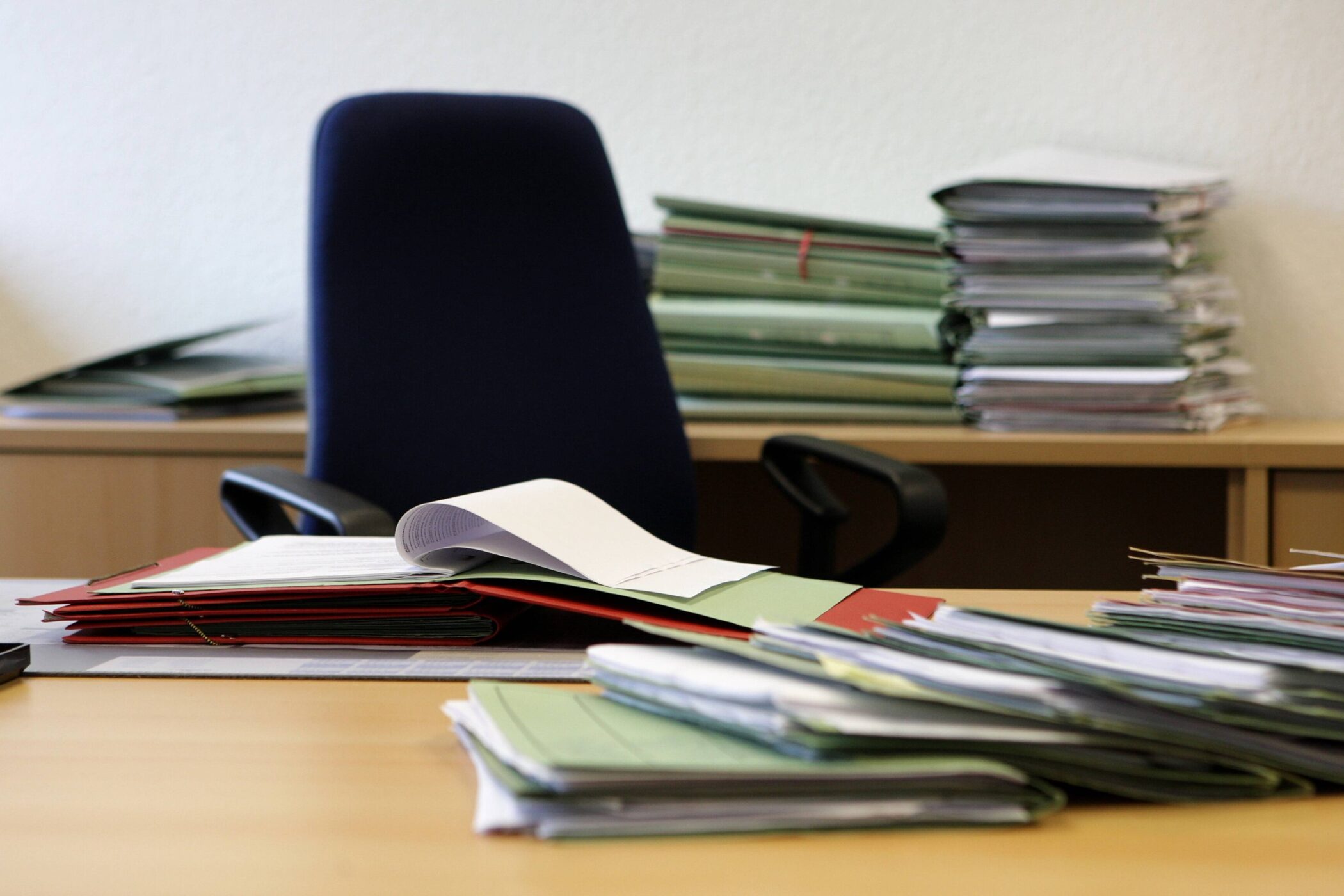 Schreibtisch mit Aktenstapeln und leerem Bürostuhl im Hintergrund.