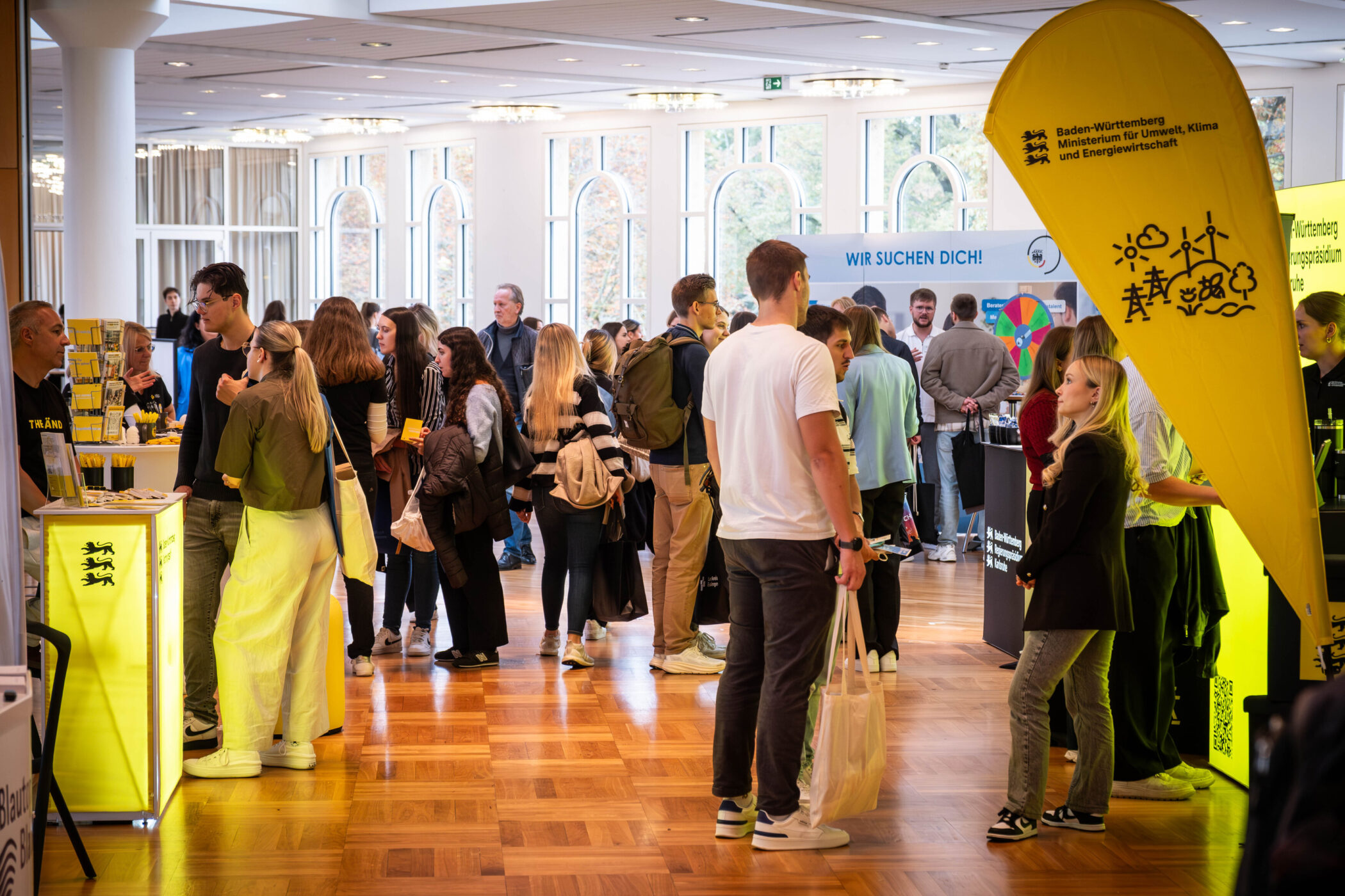 Menschenmenge bei einer Messe mit gelbem Banner und Infoständen.
