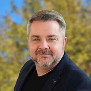 Ein Mann mit Bart lächelt vor unscharfem Hintergrund mit Bäumen.