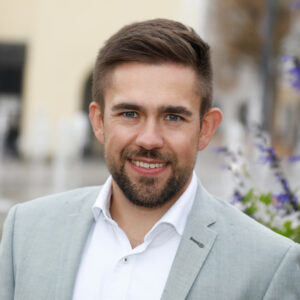 Ein Mann in hellem Anzug lächelt in die Kamera, im Hintergrund Pflanzen.