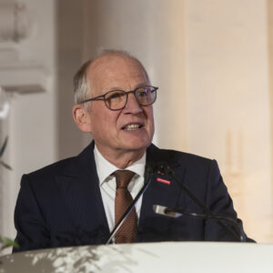 Älterer Mann mit Brille spricht am Rednerpult, Blumen im Hintergrund.