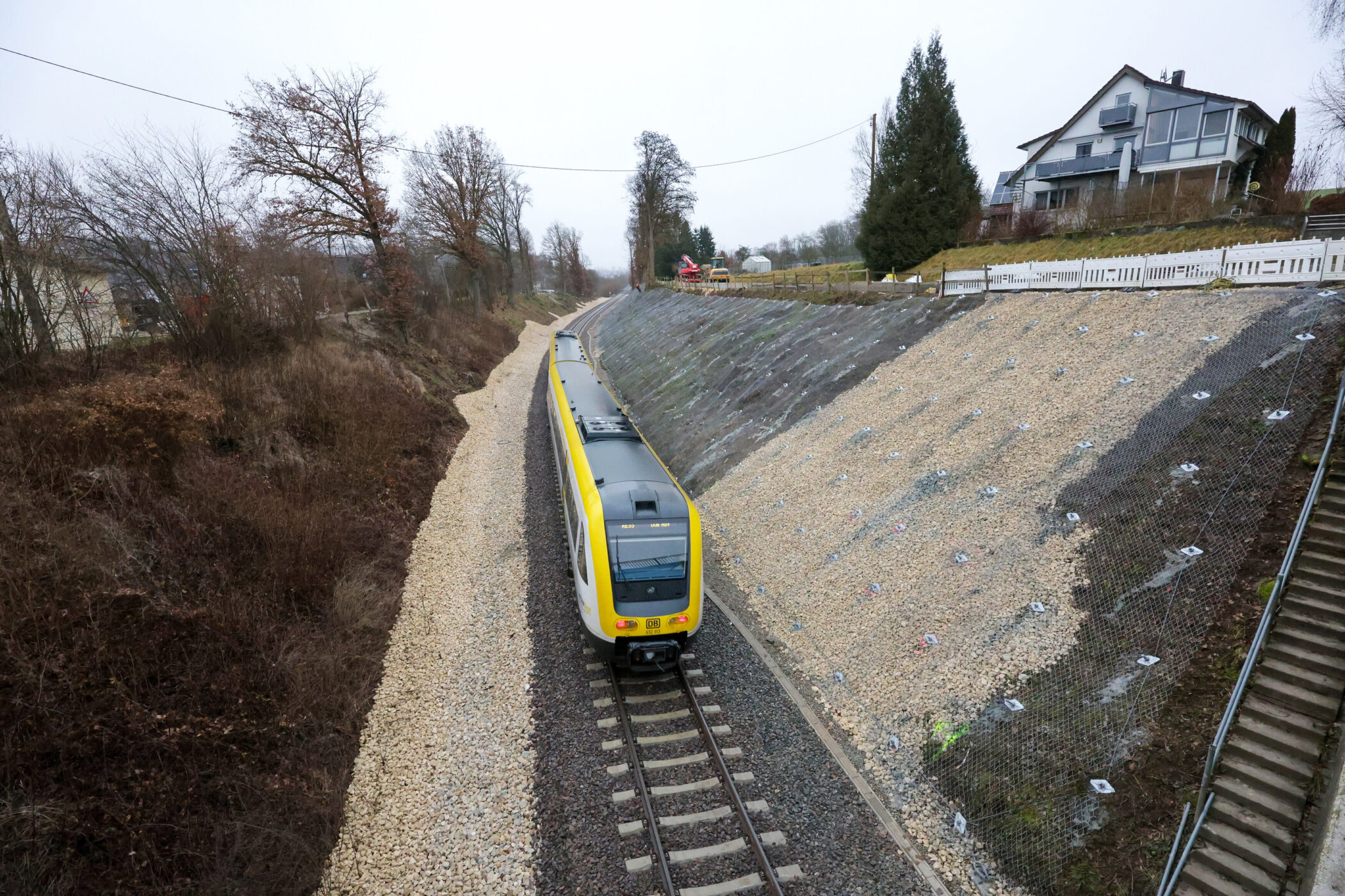 Zug auf Gleis neben steiler Böschung, Häuser und Bäume im Hintergrund.