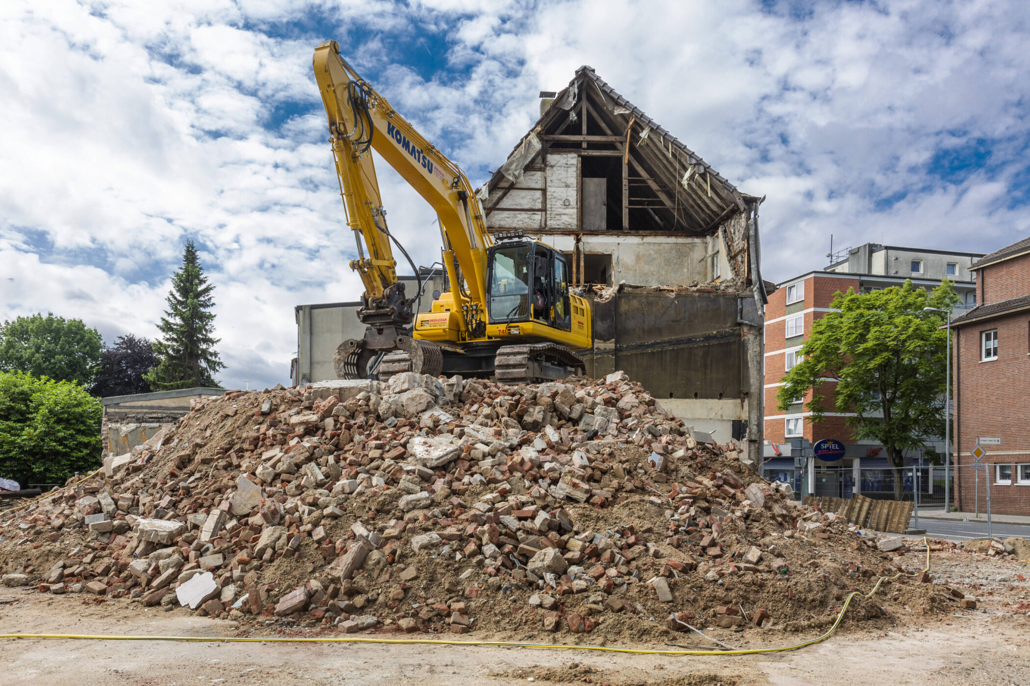 Bagger auf Schutthaufen vor halb abgerissenem Gebäude, blauer Himmel im Hintergrund.