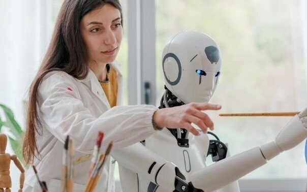 Frau in weißem Kittel zeigt einem humanoiden Roboter mit Pinsel etwas.