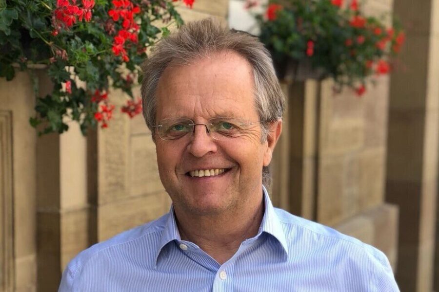 Ein lächelnder Mann mit Brille vor einer Wand mit roten Blumen.