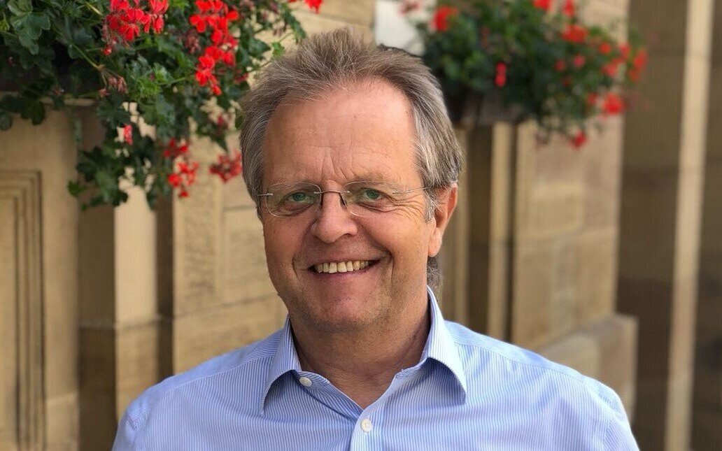 Ein lächelnder Mann mit Brille vor einer Wand mit roten Blumen.