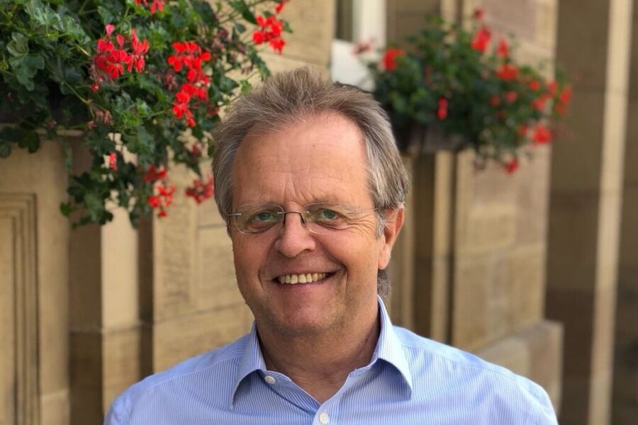 Mann mit Brille lächelt, im Hintergrund rote Blumen an einer Hauswand.