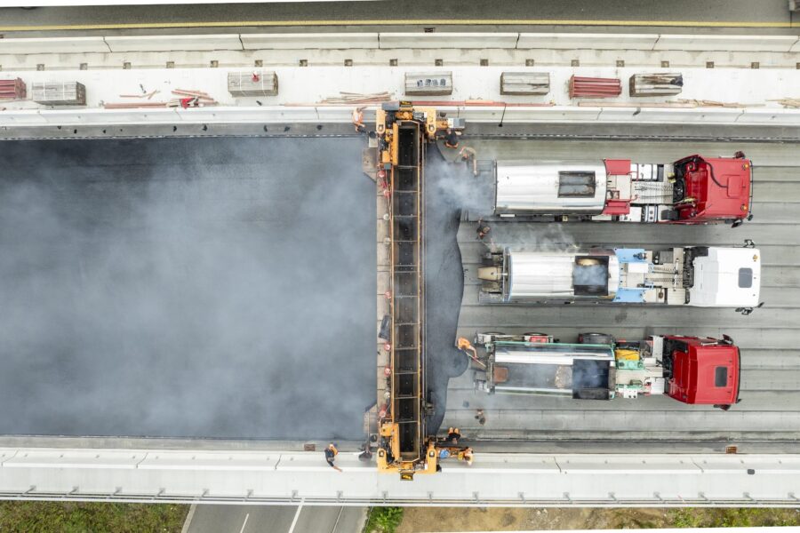 Drei Lastwagen und Arbeiter auf einer Baustelle, Asphalt wird aufgetragen.