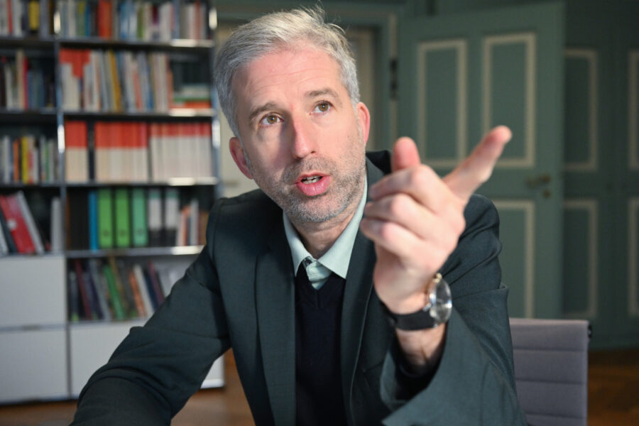 Mann in Anzug gestikuliert, Bücherregal im Hintergrund.