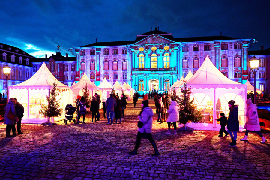 Menschen auf einem beleuchteten Weihnachtsmarkt mit weißen Zelten und einem großen Gebäude.
