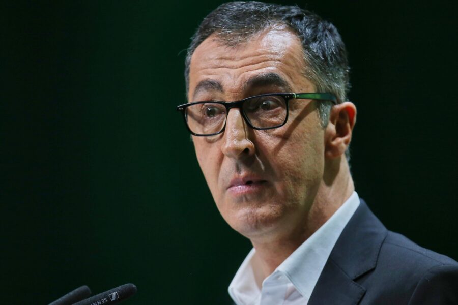 Mann mit Brille spricht in ein Mikrofon, trägt Anzug und weißes Hemd.