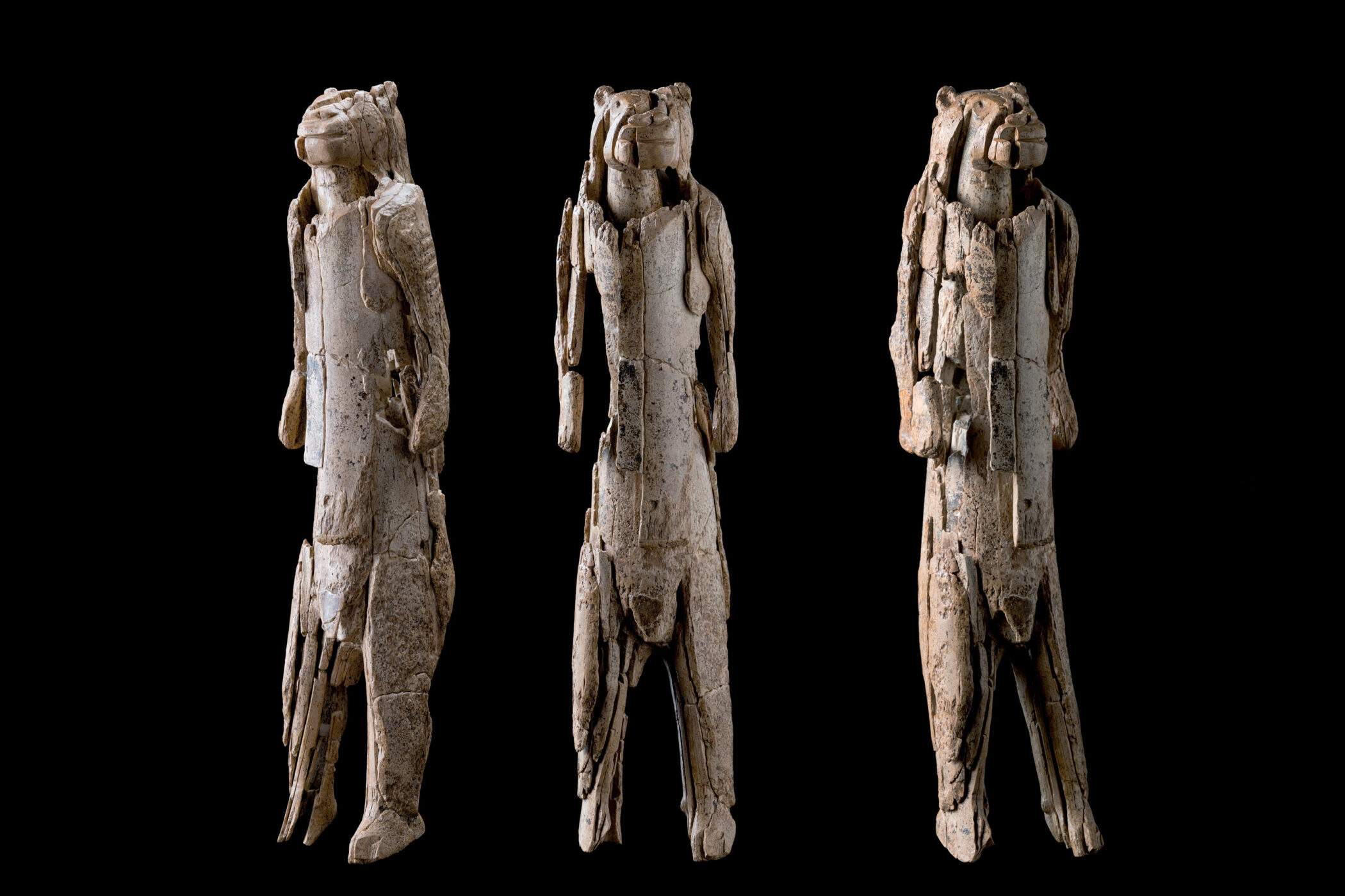 Drei geschnitzte Figuren mit tierähnlichen Köpfen auf schwarzem Hintergrund.