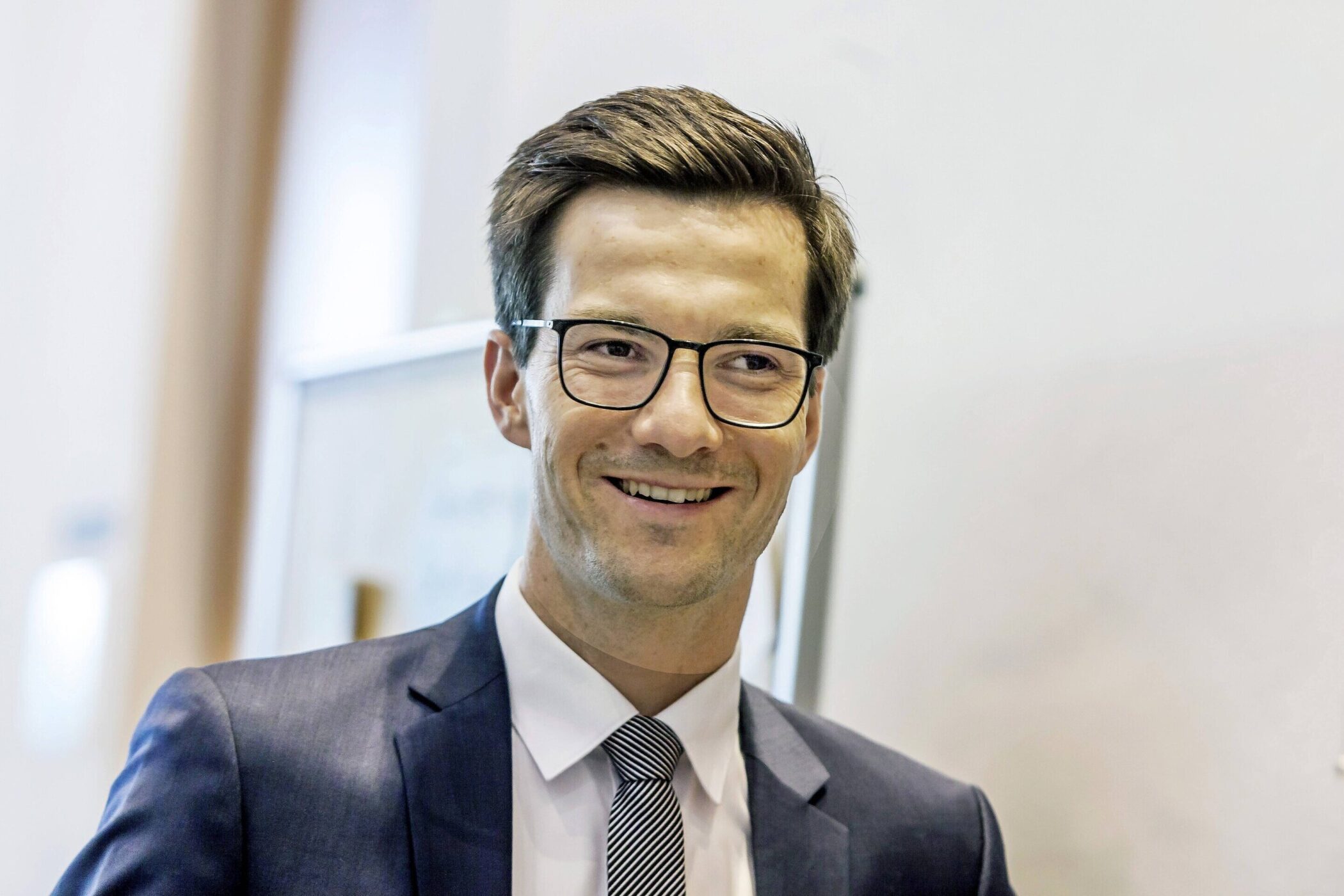 Mann mit Brille und Anzug lächelt, Hintergrund unscharf.