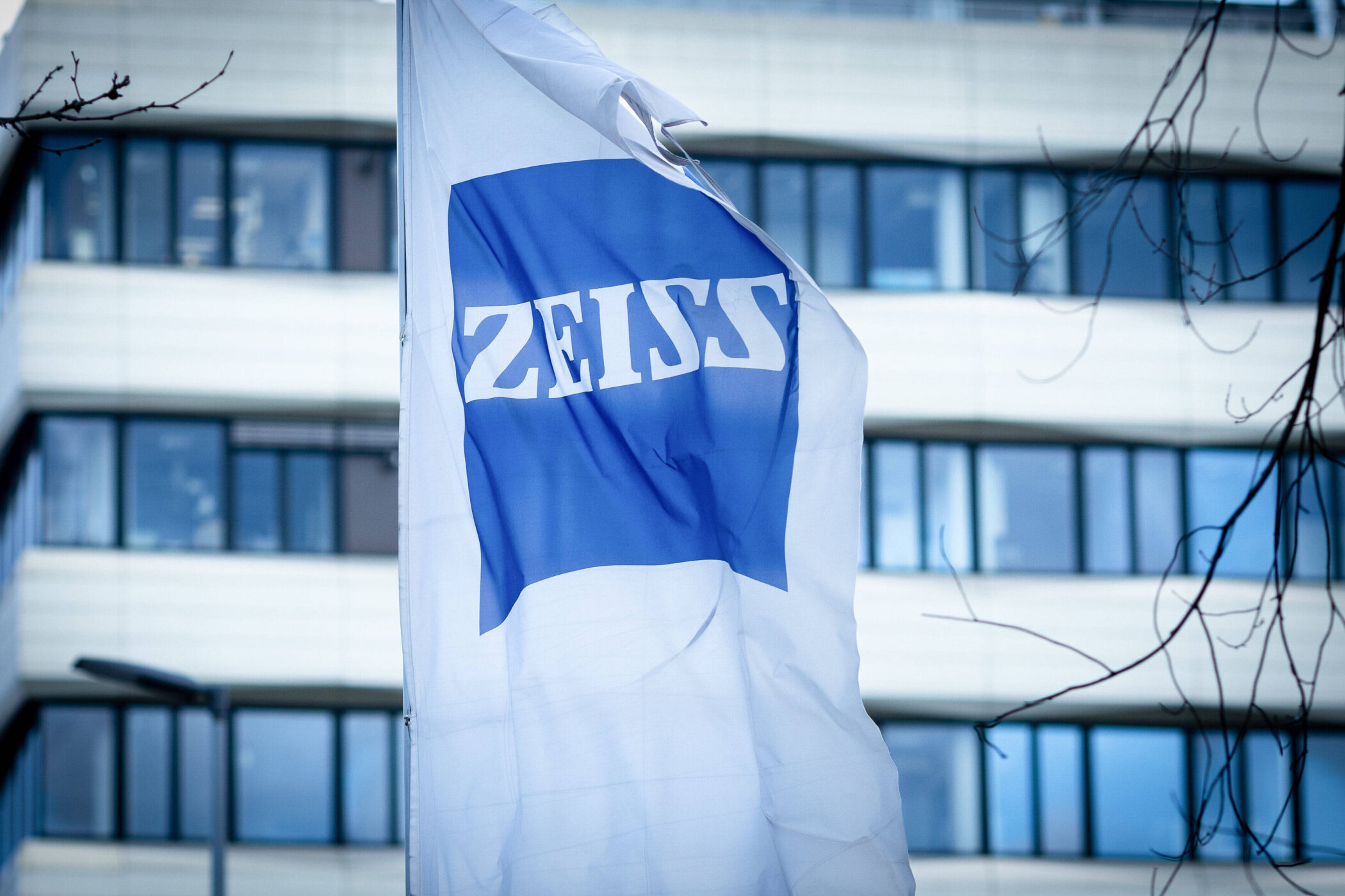 Weiße Fahne mit blauem "ZEISS"-Logo vor einem Gebäude mit Fenstern.