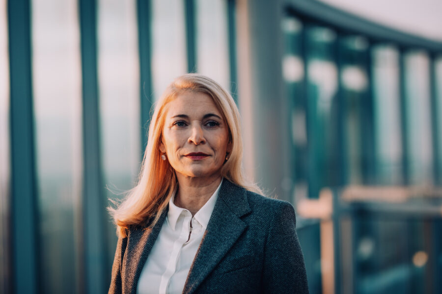 Frau mit blonden Haaren in Anzug vor Glasgebäude.