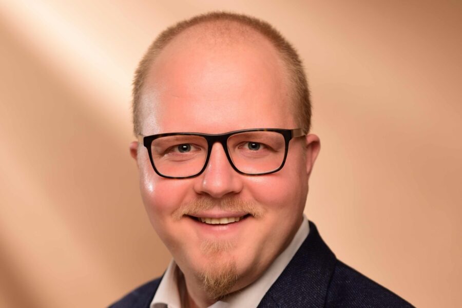 Mann mit Brille und Bart lächelt vor beigem Hintergrund.