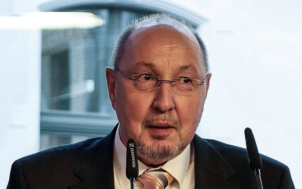 Älterer Mann mit Brille und Anzug spricht in Mikrofonen.