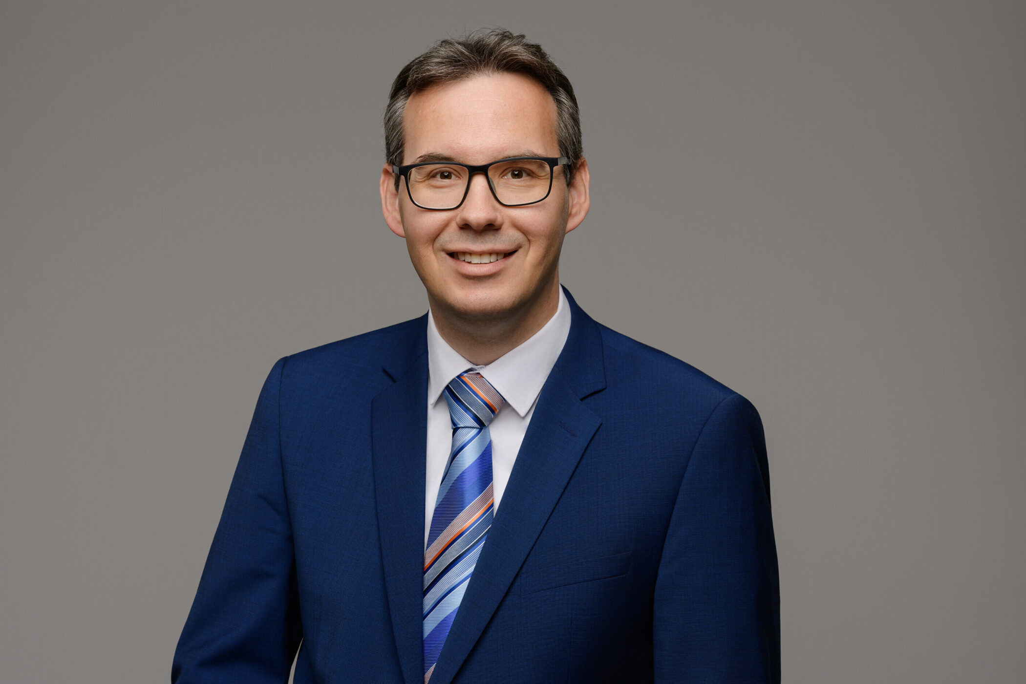 Mann mit Brille im blauen Anzug und gestreifter Krawatte vor grauem Hintergrund.
