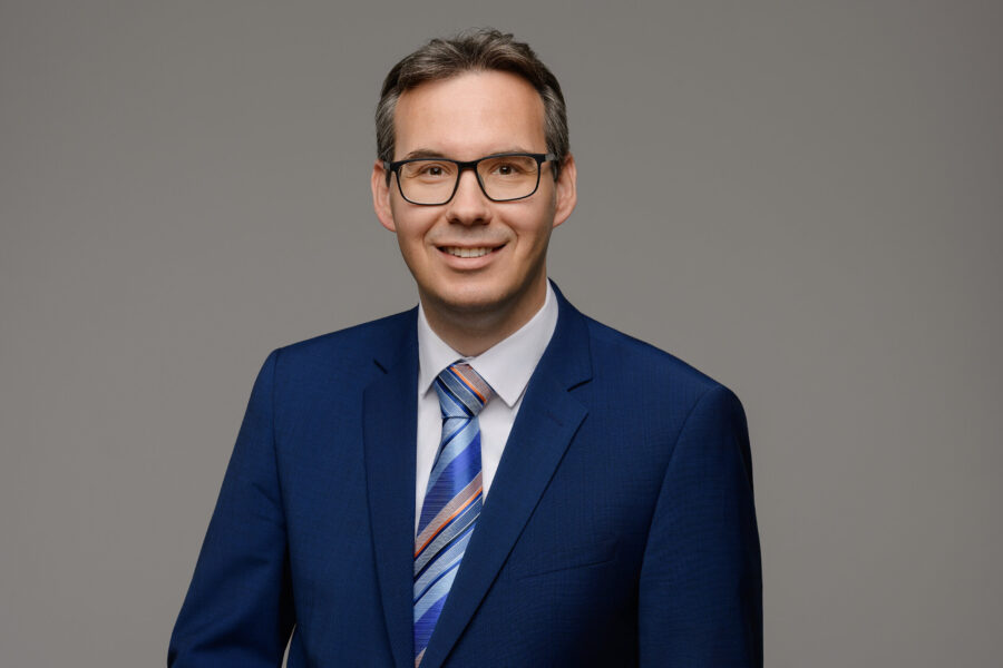 Mann mit Brille im blauen Anzug und gestreifter Krawatte vor grauem Hintergrund.