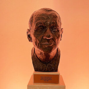 Bronzebüste eines Mannes auf Sockel mit Inschrift. Hintergrund orange beleuchtet.