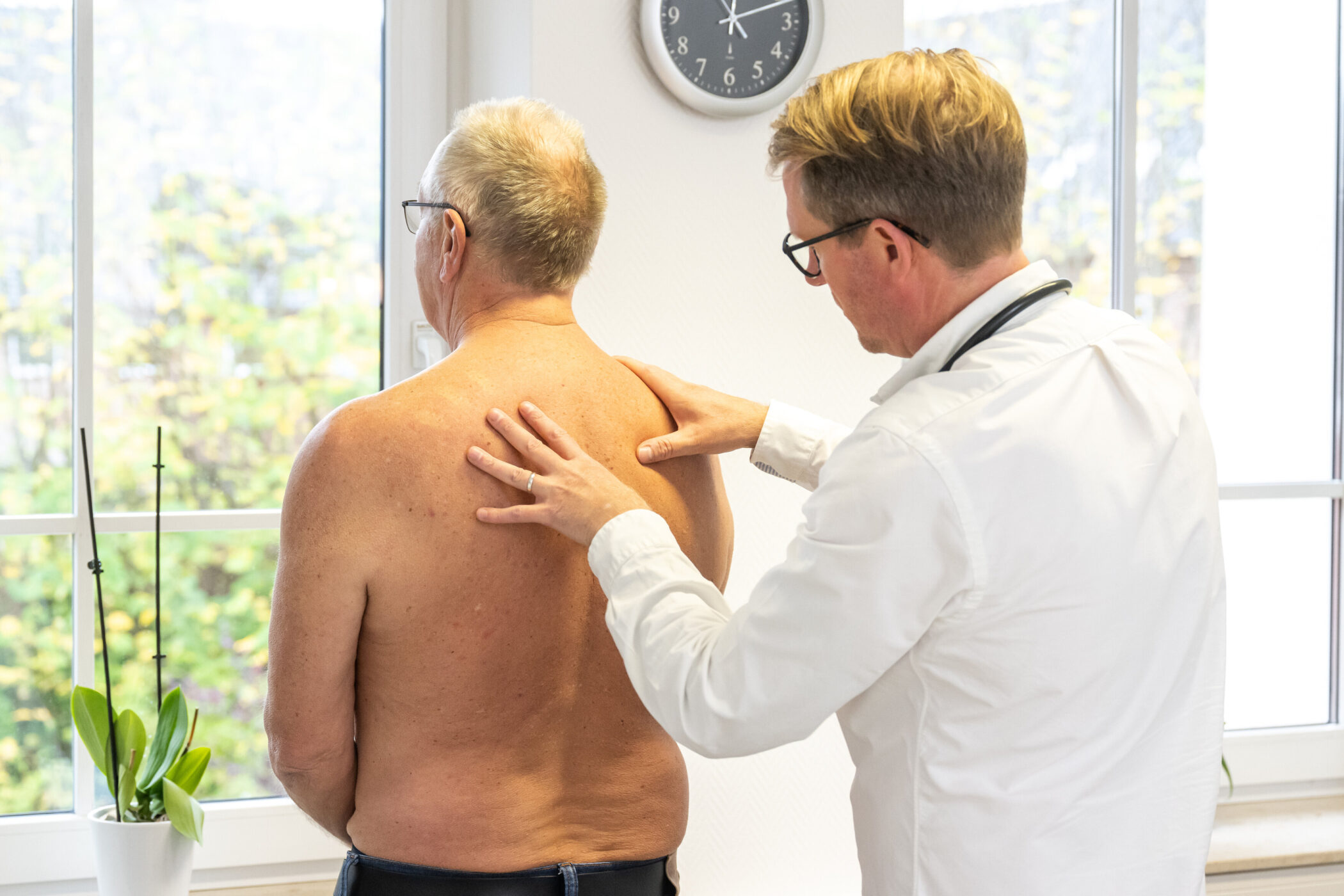 Zwei Männer, einer untersucht den Rücken des anderen, im Raum mit Fenster und Uhr.