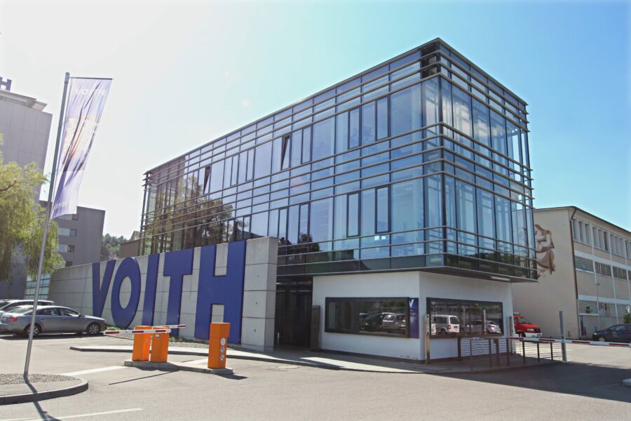 Modernes Gebäude mit Glasfassade, großer Schriftzug "VOITH", Parkplatz im Vordergrund.