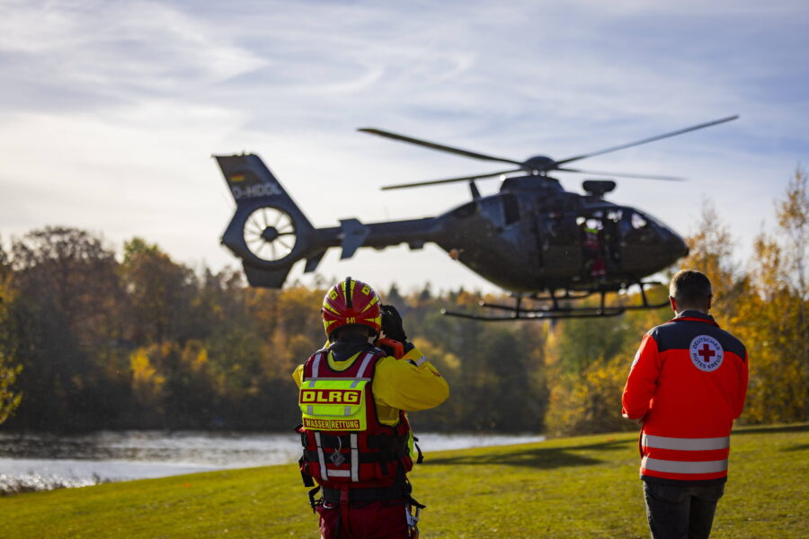 Zwei Personen in Rettungswesten beobachten einen Helikopter über einer Wiese.