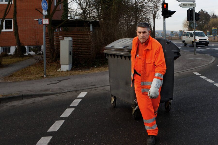 Person in orange Arbeitskleidung zieht Müllcontainer über Straße.