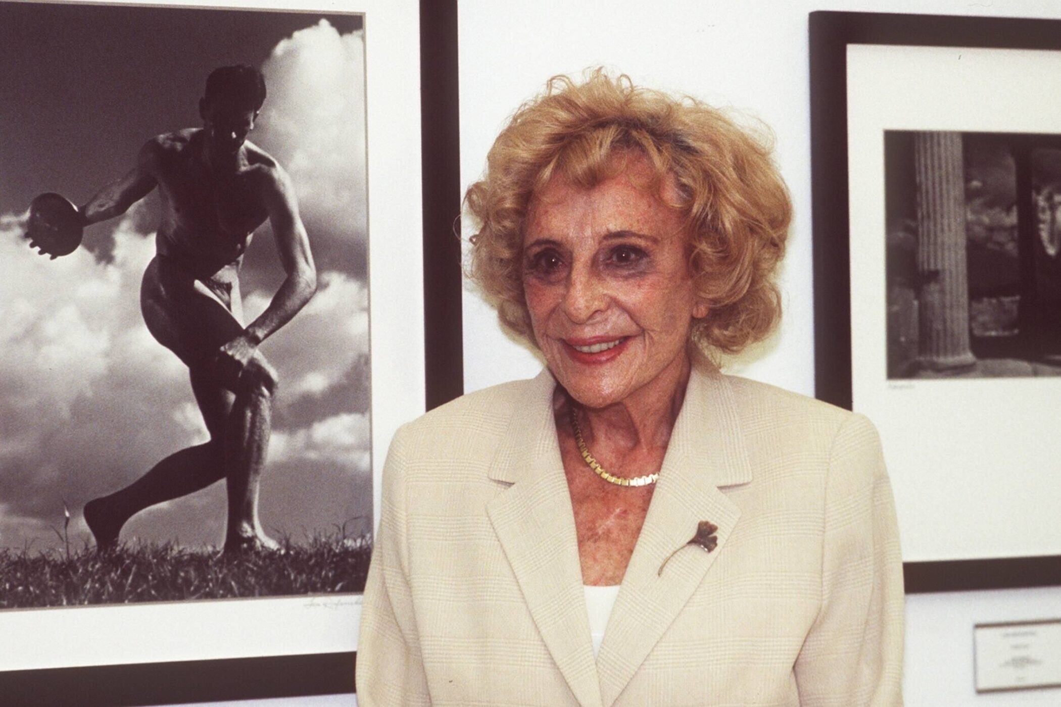 Ältere Frau in hellem Blazer vor zwei gerahmten Schwarz-Weiß-Fotos an der Wand.