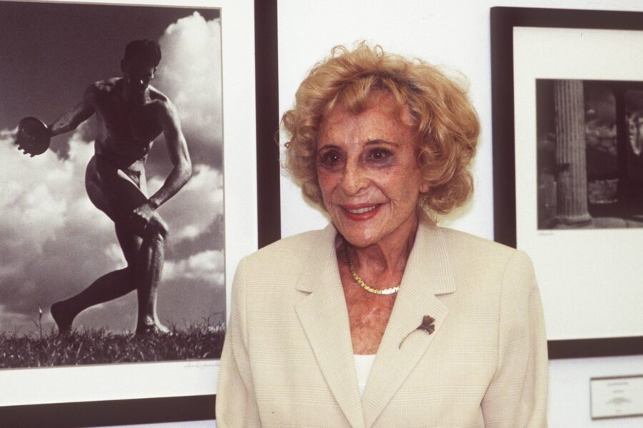 Ältere Frau in hellem Blazer vor zwei gerahmten Schwarz-Weiß-Fotos an der Wand.