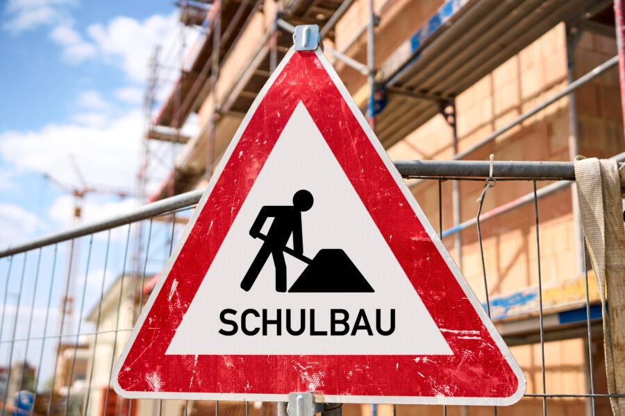 Dreieckiges Schild mit Bauarbeiter-Symbol und Text "SCHULBAU". Hintergrund: Baustelle.