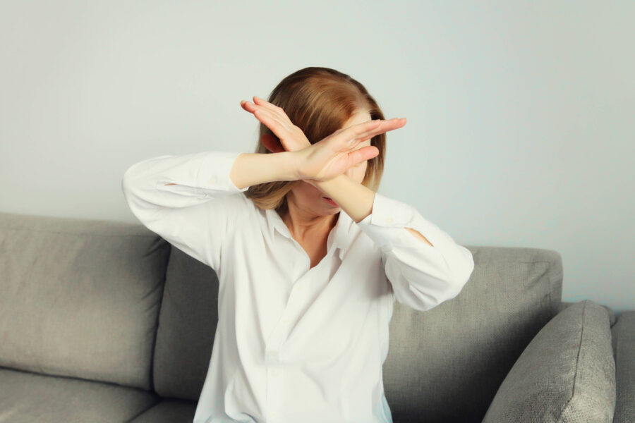 Person auf Sofa, hält Arme vor Gesicht gekreuzt, trägt weißes Hemd.