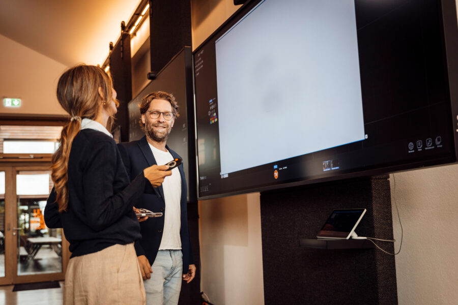 Zwei Personen vor einem großen Bildschirm, eine hält eine Fernbedienung.