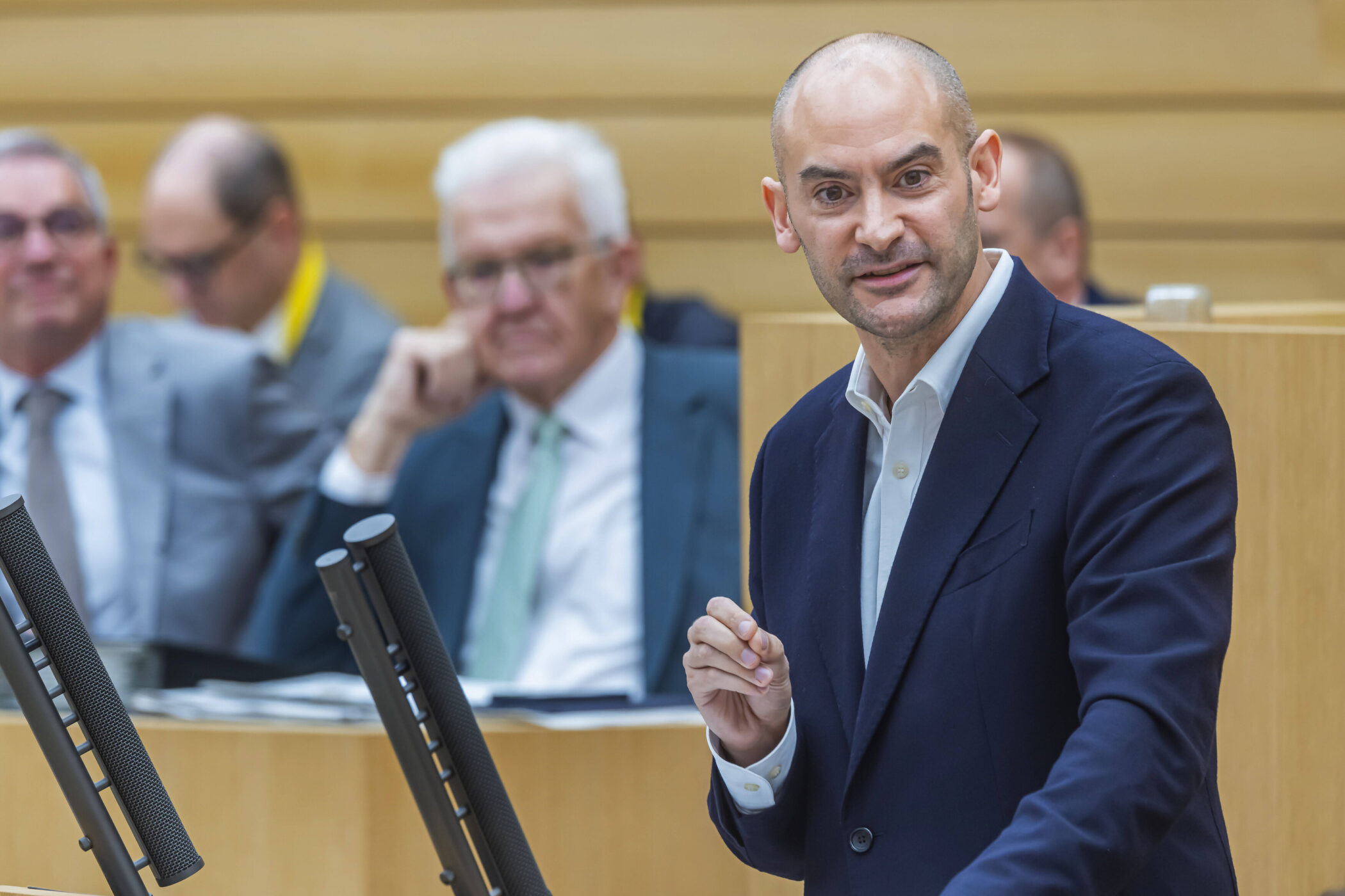 Ein Mann in Anzug spricht an einem Rednerpult, mehrere Männer im Hintergrund.