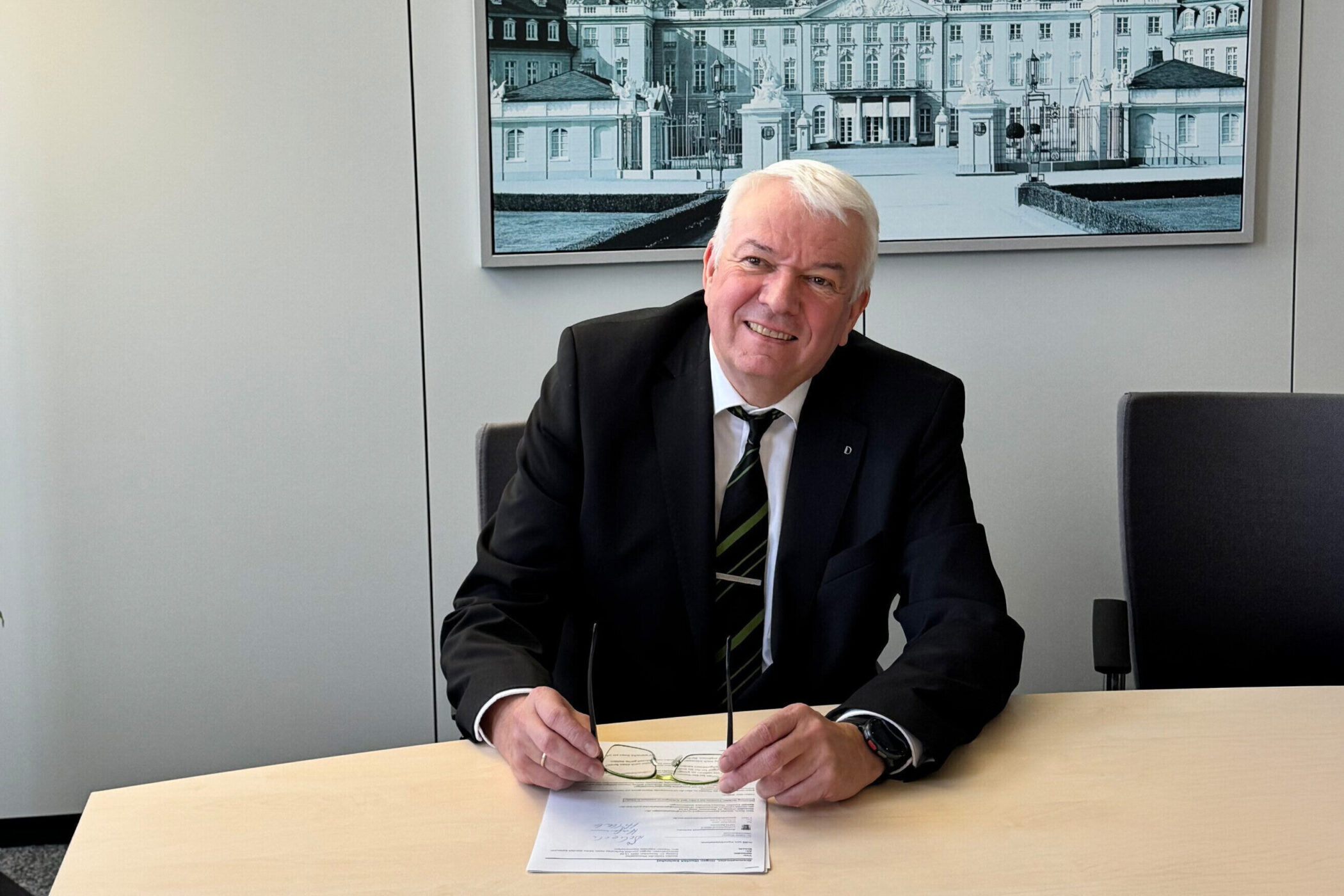 Älterer Mann im Anzug sitzt an einem Tisch, hält eine Brille, Bild im Hintergrund.