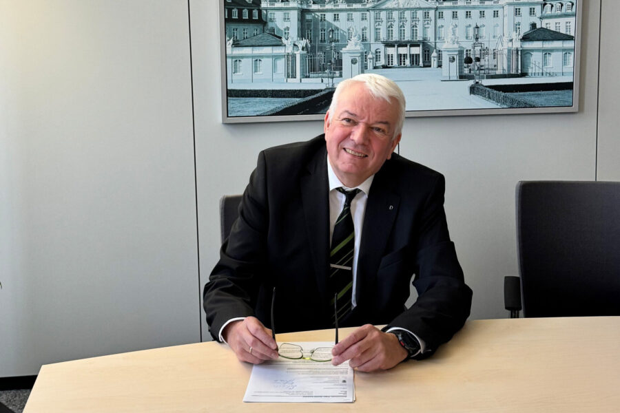 Älterer Mann im Anzug sitzt an einem Tisch, hält eine Brille, Bild im Hintergrund.